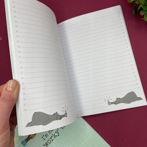 May include: Open notebook with lined pages and check boxes. The notebook features a light green cover with a grid pattern and the text "I'm not very 'worky'". Inside, a cartoon bear is drawn on the bottom of the pages, holding a mug.