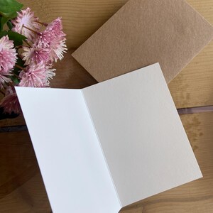 May include: A blank white card with a brown envelope. The card is folded in half and has a smooth, textured surface.