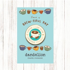 May include: A light blue card with the text "Have a BREW-TIFUL DAY" and a "Coffee Lover Club Lifetime Member" enamel pin badge. The badge features a smiling coffee cup. The card also has the word "dandelion" and "ENAMEL PIN BADGE".