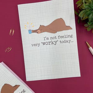 May include: A light blue greeting card with a brown bear illustration holding a mug. The text reads, "I'm not feeling very 'worky' today..." The card has a grid pattern and is on a burgundy surface.