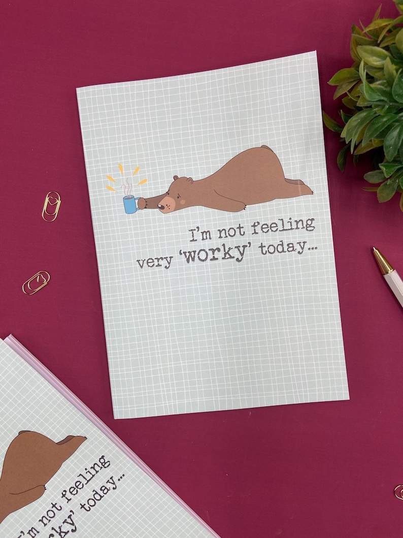 May include: A light blue greeting card with a brown bear illustration holding a mug. The text reads, "I'm not feeling very 'worky' today..." The card has a grid pattern and is on a burgundy surface.