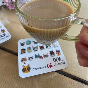 May include: A white coaster with a colorful illustration of cats wearing different hats and accessories. The text "OCD... Obsessive Cat Disorder" is printed on the coaster.