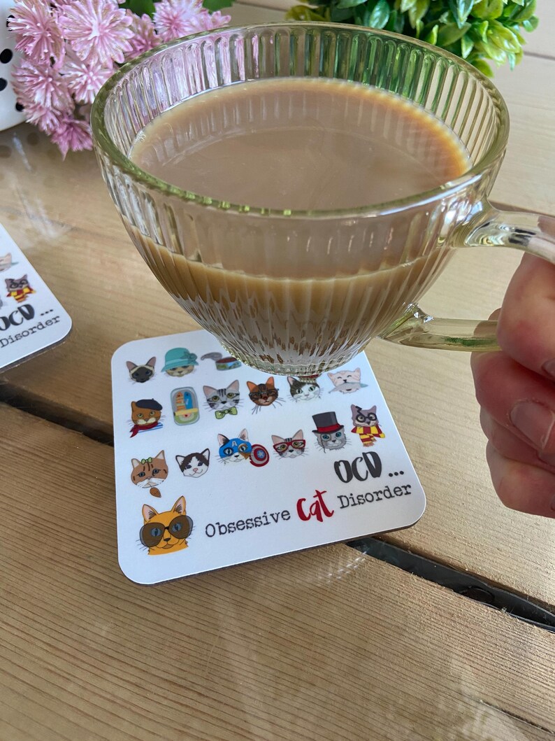 May include: A white coaster with a colorful illustration of cats wearing different hats and accessories. The text "OCD... Obsessive Cat Disorder" is printed on the coaster.