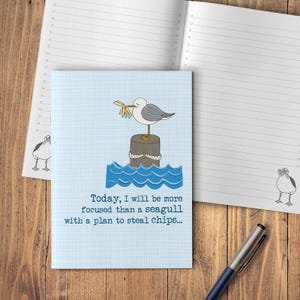 May include: A blue notebook featuring a seagull illustration. The seagull is depicted on a wooden post, holding food. The notebook has lined pages and the text: "Today, I will be more focused than a seagull with a plan to steal chips..."