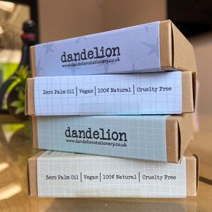 May include: Four stacked boxes of stationery products. The boxes are light brown with pastel-colored labels. The labels feature the brand name "dandelion" and the website address. The labels also state "Zero Palm Oil, Vegan, 100% Natural, Cruelty Free."