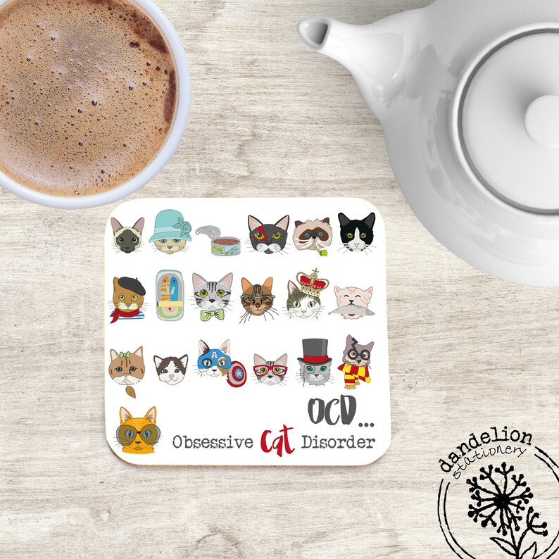 May include: A white square coaster with a humorous design featuring a grid of 20 different cat illustrations wearing various hats, glasses, and accessories. The text "OCD... Obsessive Cat Disorder" is printed below the grid.