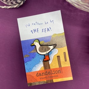 May include: A colourful enamel pin badge of a seagull wearing a green hat and red wellington boots, perched on a wooden post. The card reads "I'd rather be by THE SEA!" and features the brand name "dandelion".