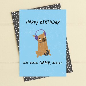 May include: A blue birthday card featuring a cartoon dog wearing headphones and holding a video game controller. The text on the card reads "Happy Birthday" and "Eat, Sleep, Game, Repeat."