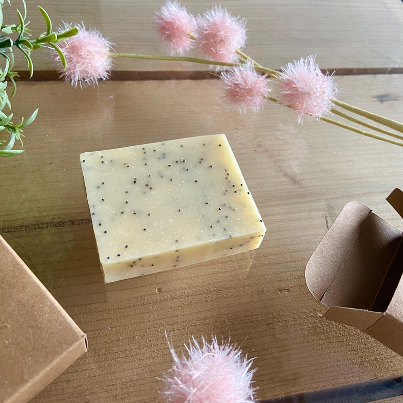 May include: A square bar of handmade soap with black specks, likely poppy seeds, on a light yellow background. The soap is displayed on a wooden surface with pink decorative flowers and a small brown cardboard box.