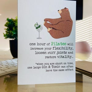 May include: A humorous greeting card featuring a cartoon bear doing a yoga pose with a glass of gin and tonic next to it. The card reads "one hour of Pilates will increase your flexibility, loosen stiff joints and restore vitality... *when you are short on time, one large Gin & Tonic can often have the same effect"