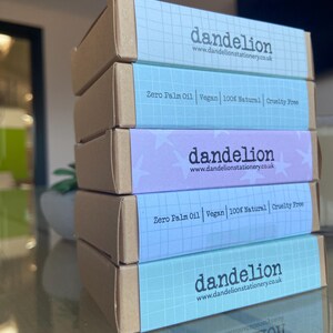 May include: A stack of five rectangular boxes with brown sides and pastel blue, purple, and teal tops. The boxes feature the word "dandelion" and the website address www.dandelionstationery.co.uk. The boxes also state "Zero Palm Oil Vegan 100% Natural Cruelty Free."