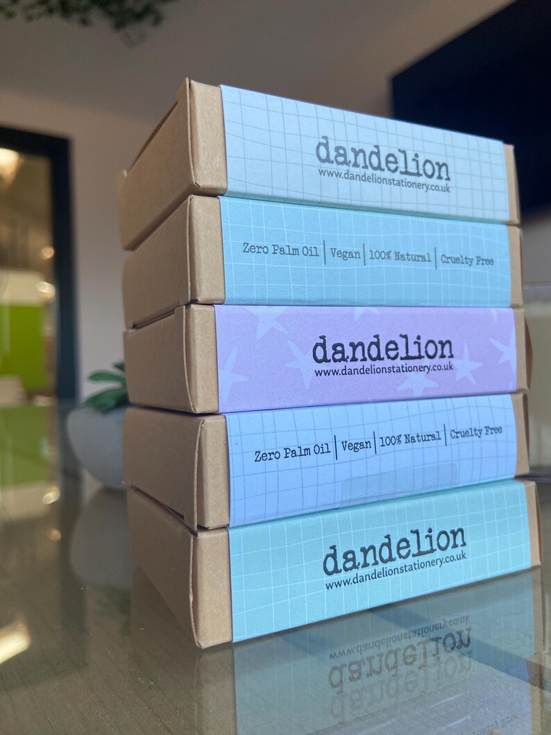 May include: A stack of five rectangular boxes with brown sides and pastel blue, purple, and teal tops. The boxes feature the word "dandelion" and the website address www.dandelionstationery.co.uk. The boxes also state "Zero Palm Oil Vegan 100% Natural Cruelty Free."