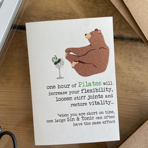 May include: A humorous greeting card with a cartoon illustration of a brown bear doing a yoga pose. The text reads: "One hour of Pilates will increase your flexibility, loosen stiff joints and restore vitality... *when you are short on time, one large Gin & Tonic can often have the same effect"