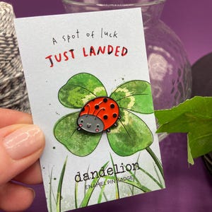 May include: A ladybird enamel pin badge on a card with the text "A spot of luck JUST LANDED". The pin features a red ladybird with black spots on a green four-leaf clover. The card also says "dandelion ENAMEL PIN BADGE".