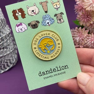 May include: A light green card with cartoon dog illustrations and a gold enamel pin. The pin says "DOG LOVER CLUB LIFETIME MEMBER" and features a yellow dog design. The card also has the text "dandelion ENAMEL PIN BADGE".