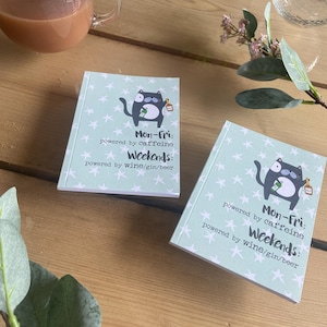 May include: Two small, square notebooks with a light blue background and white star pattern. Each notebook features a cartoon cat graphic and text that reads "Mon-Fri: powered by caffeine, Weekends: powered by wine/gin/beer."