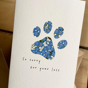 May include: A white sympathy card featuring a paw print design filled with blue and white floral patterns. The words "So sorry for your loss" are written in black script below the paw print. The card is held in front of a brown envelope.