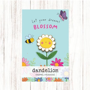 May include: A light blue enamel pin badge with a dandelion design. The pin features a yellow flower with a smiling face, a bee, and a butterfly. The text "Let your dreams BLOSSOM" is above the flower, and "dandelion ENAMEL PIN BADGE" is below.