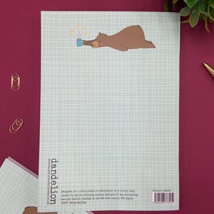 May include: A light blue notepad with a grid pattern and a brown bear illustration holding a blue mug with steam. The notepad has the text "dandelion" and other text. The notepad is on a burgundy surface.