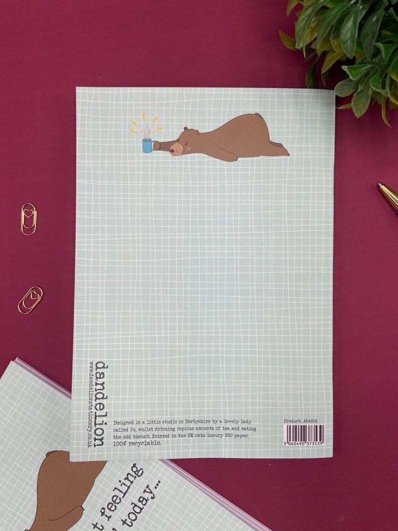 May include: A light blue notepad with a grid pattern and a brown bear illustration holding a blue mug with steam. The notepad has the text "dandelion" and other text. The notepad is on a burgundy surface.