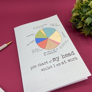 Puede incluir: Un cuaderno azul claro con un gr&aacute;fico de tarta titulado "pie chart of my head whilst I am at work". El gr&aacute;fico incluye frases humor&iacute;sticas relacionadas con el trabajo. Un bol&iacute;grafo es visible en el fondo.