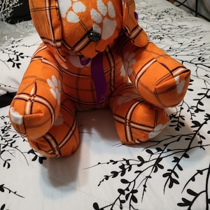 May include: Orange and purple plaid stuffed dog with white paw prints. The dog is sitting on a white bedspread with a black floral pattern.