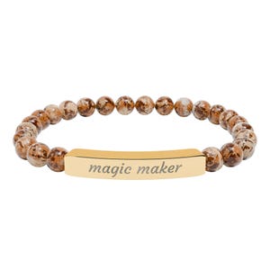 Può includere: Bracciale con perline maculate marroni e beige. Una barra rettangolare color oro al centro reca la scritta "magic maker" in corsivo. Il bracciale è infilato su un elastico.