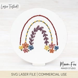 May include: A white, round wooden embroidery hoop featuring a colorful embroidered rainbow design. The rainbow has a dark red outer arc, with floral and leaf accents in shades of yellow, purple, orange, and blue. Text at the top reads "Laser Tested!"