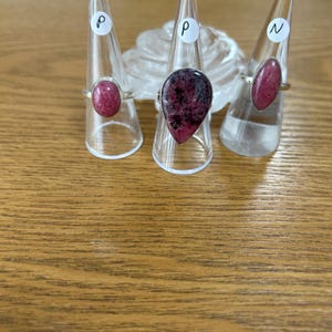 May include: Three rings with oval and teardrop-shaped gemstones are displayed on clear plastic stands. The gemstones are shades of pink and red, set in silver bands. The rings are arranged on a wooden surface.
