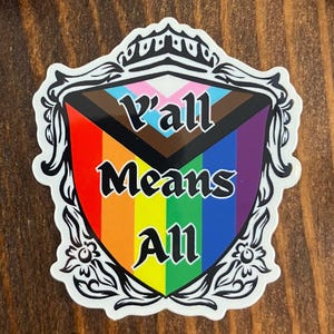 May include: A white sticker with a shield design featuring the text "Y'all Means All" in black. The shield incorporates the colors of the rainbow and the transgender flag. The sticker is surrounded by a decorative black border.