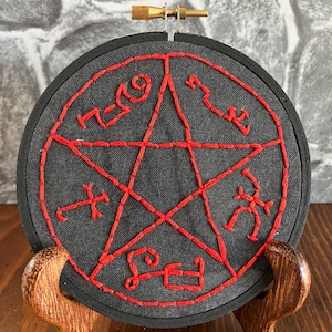 May include: A black embroidery hoop featuring a red pentagram design with symbols. The embroidery is mounted on a wooden stand. The design is stitched with red thread on a black fabric.