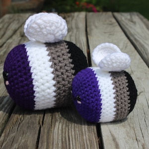 May include: Two crocheted bee plushies, one larger than the other, with purple, white, gray, and black stripes. The bees have white crocheted wings.