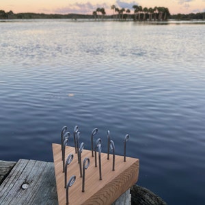 Può includere: Un pannello triangolare di legno con ganci metallici che sporgono dalla superficie. Il pannello è appoggiato su un molo di legno che si affaccia su uno specchio d'acqua.