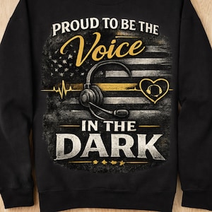 May include: A black sweatshirt featuring a graphic design with the text "PROUD TO BE THE VOICE IN THE DARK". The design includes a stylized American flag, a headset, a heartbeat line, and a heart symbol in yellow and white on a black background.