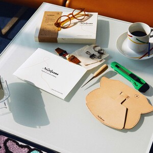 May include: A white table with a variety of leatherworking tools and supplies, including a leather pouch, a small awl, a craft knife, a pair of glasses, a small box, a cup of coffee, and a small fan. The table is covered in a white tablecloth. The text "Artisans" is visible on the box and the leather pouch.