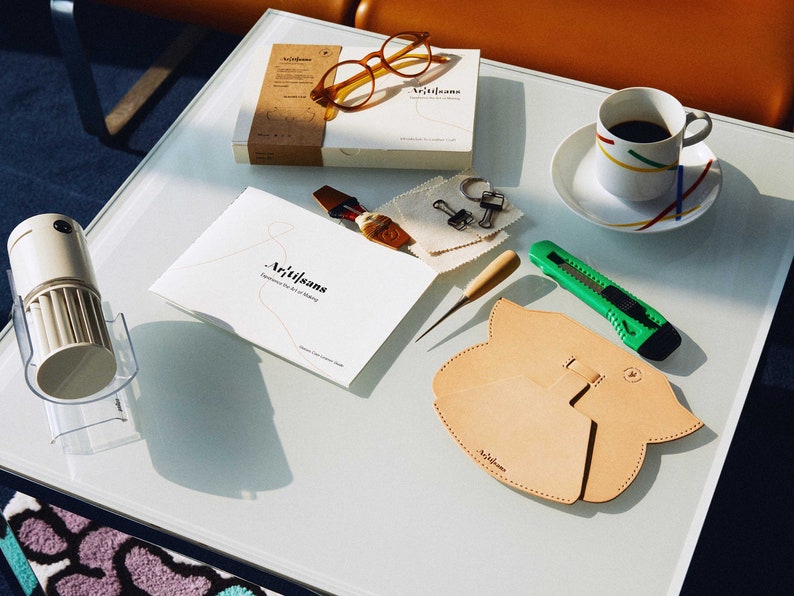 May include: A white table with a variety of leatherworking tools and supplies, including a leather pouch, a small awl, a craft knife, a pair of glasses, a small box, a cup of coffee, and a small fan. The table is covered in a white tablecloth. The text "Artisans" is visible on the box and the leather pouch.