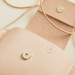 May include: A light pink leather crossbody bag with a flap closure and a gold metal snap. The bag has a small, embossed logo that reads "Designed by Crafted by Handmade with Love".