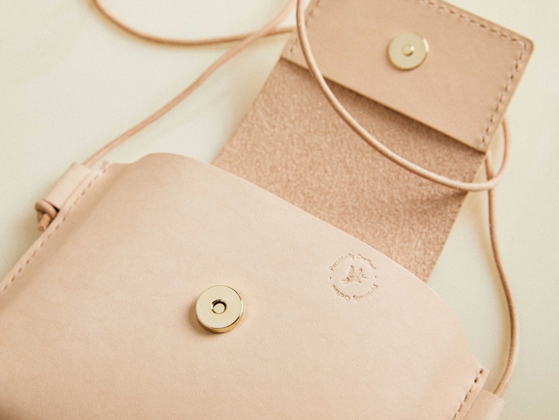 May include: A light pink leather crossbody bag with a flap closure and a gold metal snap. The bag has a small, embossed logo that reads "Designed by Crafted by Handmade with Love".