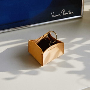 May include: A small, light brown leather key holder with the word "Artisans" embossed on the front. The holder contains a set of keys on a silver keyring. The holder is sitting on a white surface.