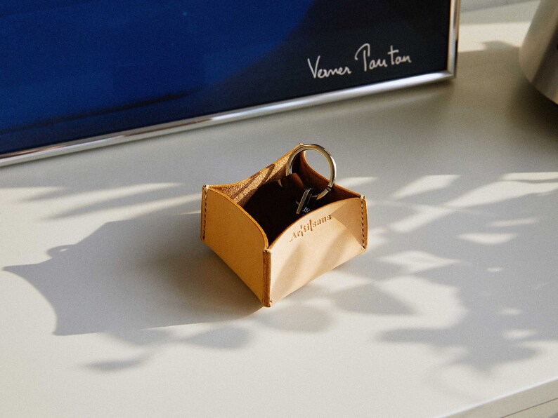 May include: A small, light brown leather key holder with the word "Artisans" embossed on the front. The holder contains a set of keys on a silver keyring. The holder is sitting on a white surface.