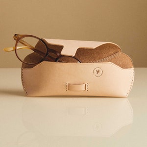 May include: A light brown leather glasses case with a flap closure and a small logo on the front. The case is open and a pair of brown glasses are inside.