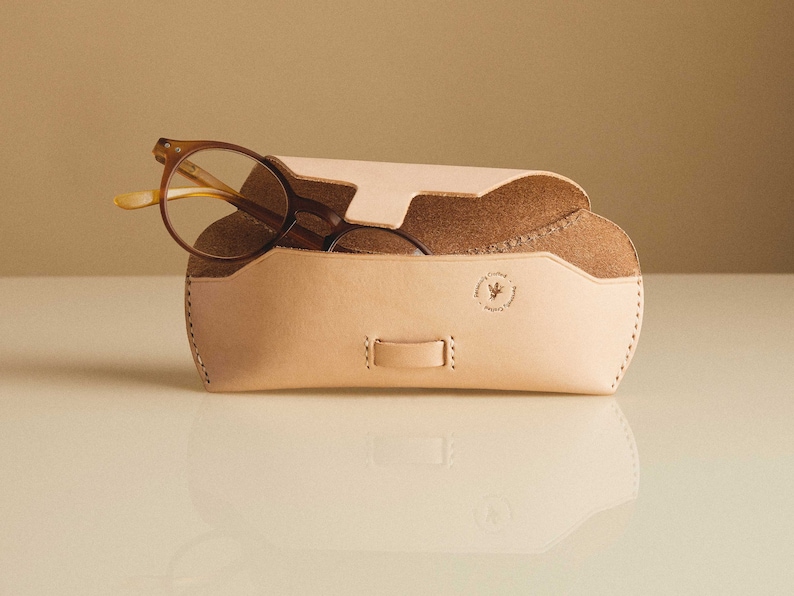 May include: A light brown leather glasses case with a flap closure and a small logo on the front. The case is open and a pair of brown glasses are inside.