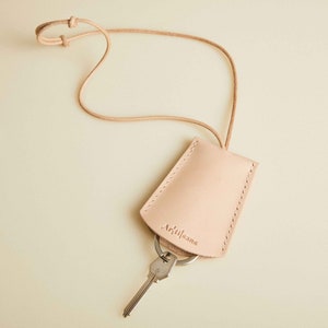 Peut inclure: Un porte-clés en cuir brun clair avec une sangle et une clé argentée attachée. Le porte-clés porte le mot "Artijeans" en relief.