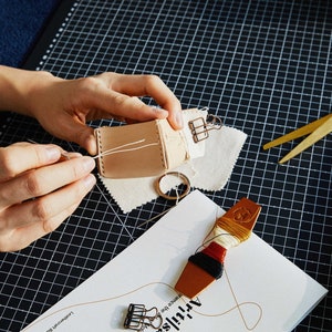 May include: A person is shown sewing a small, light brown leather pouch with white thread. The scene includes a pair of gold scissors, a spool of thread, and a paper with the word "Artisan" on it. The background is a black grid.