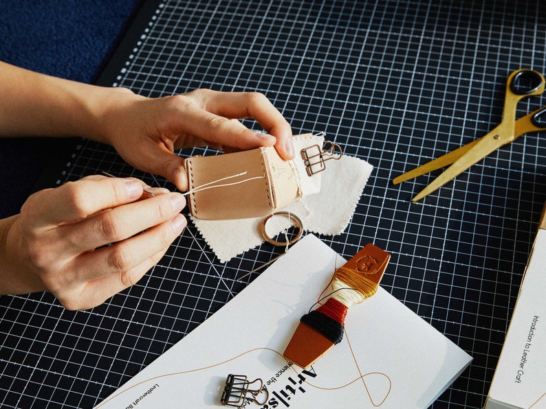 May include: A person is shown sewing a small, light brown leather pouch with white thread. The scene includes a pair of gold scissors, a spool of thread, and a paper with the word "Artisan" on it. The background is a black grid.