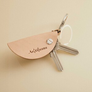 May include: A light brown leather key holder with the text "Arti|sans" embossed on the front. The key holder is attached to a silver key ring with two keys.