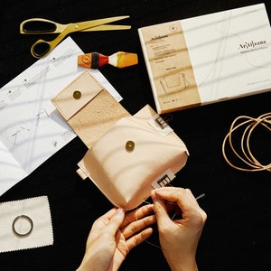 May include: A person is sewing a light brown leather pouch with a small metal button. The pouch is open and the inside is visible. The pouch is on a black surface with a white instruction sheet, a pair of scissors, a small tin, and a piece of leather cord.