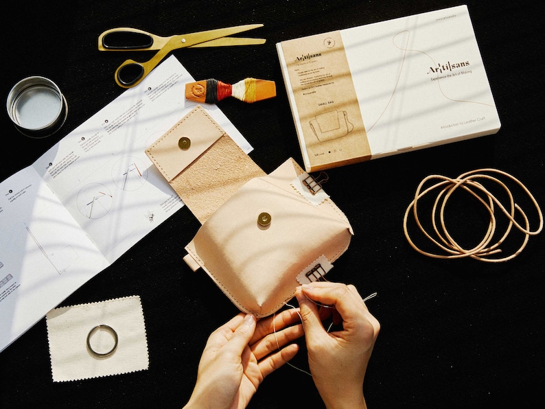 May include: A person is sewing a light brown leather pouch with a small metal button. The pouch is open and the inside is visible. The pouch is on a black surface with a white instruction sheet, a pair of scissors, a small tin, and a piece of leather cord.