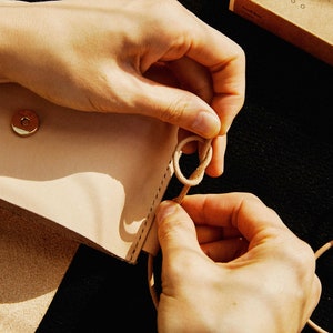 May include: A person's hands are tying a knot in a light brown leather strap. The strap is attached to a light brown leather bag with a gold metal snap closure.