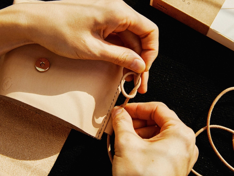 May include: A person's hands are tying a knot in a light brown leather strap. The strap is attached to a light brown leather bag with a gold metal snap closure.